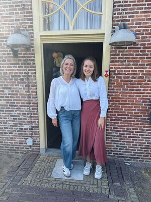 Twee vrouwen staan voor een deur, een met een witte blouse en jeans, de ander met een witte blouse en lange rok.